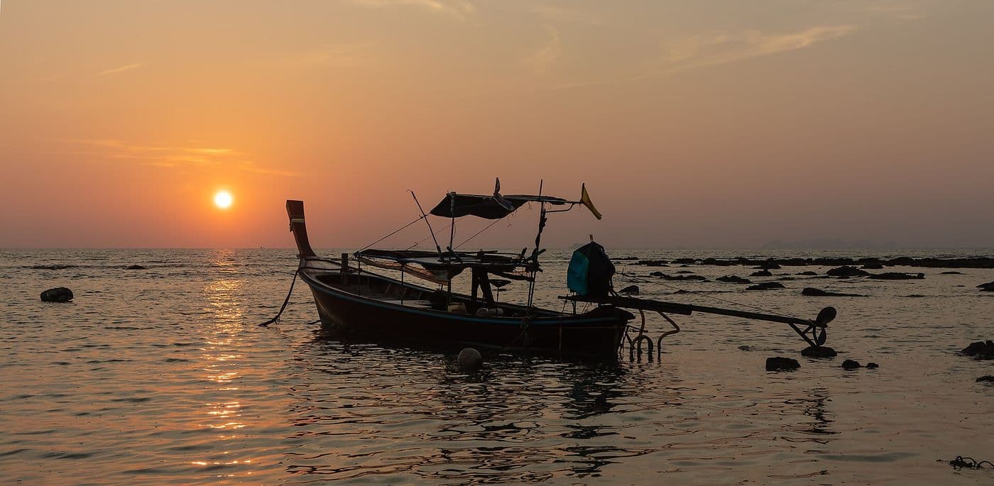 A longtail boat silhouetted on calm water at sunset off Klong Khong Beach, the sun low in an orange sky