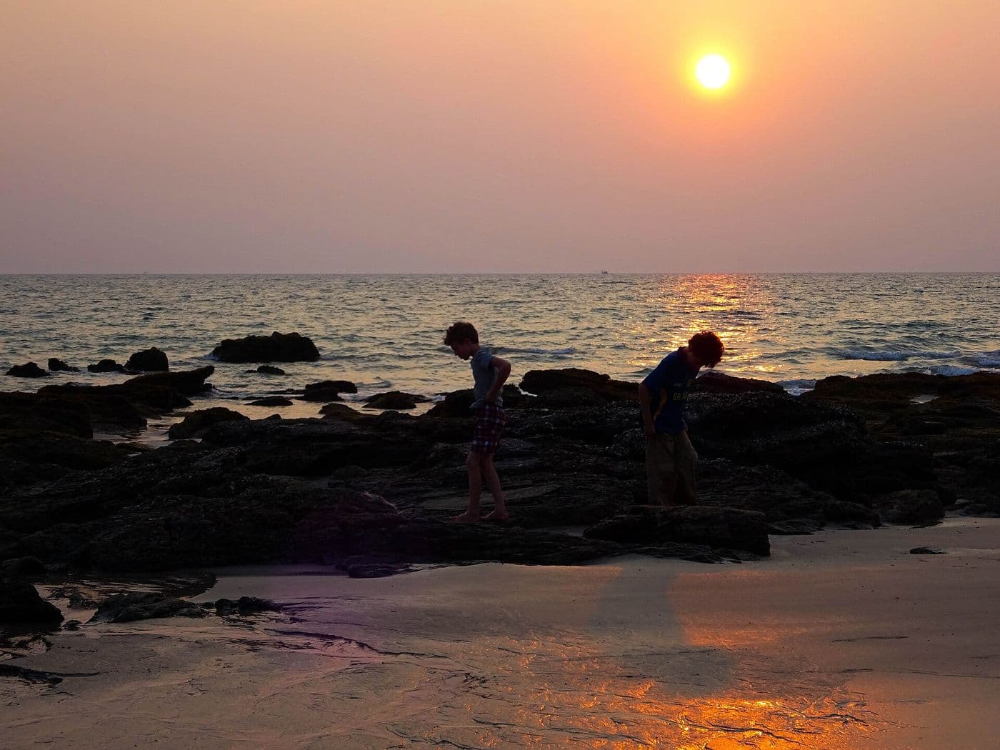 Two boys hunting for crabs among the dark rocks at low tide as the sun sets over a pink sky on Koh Lanta